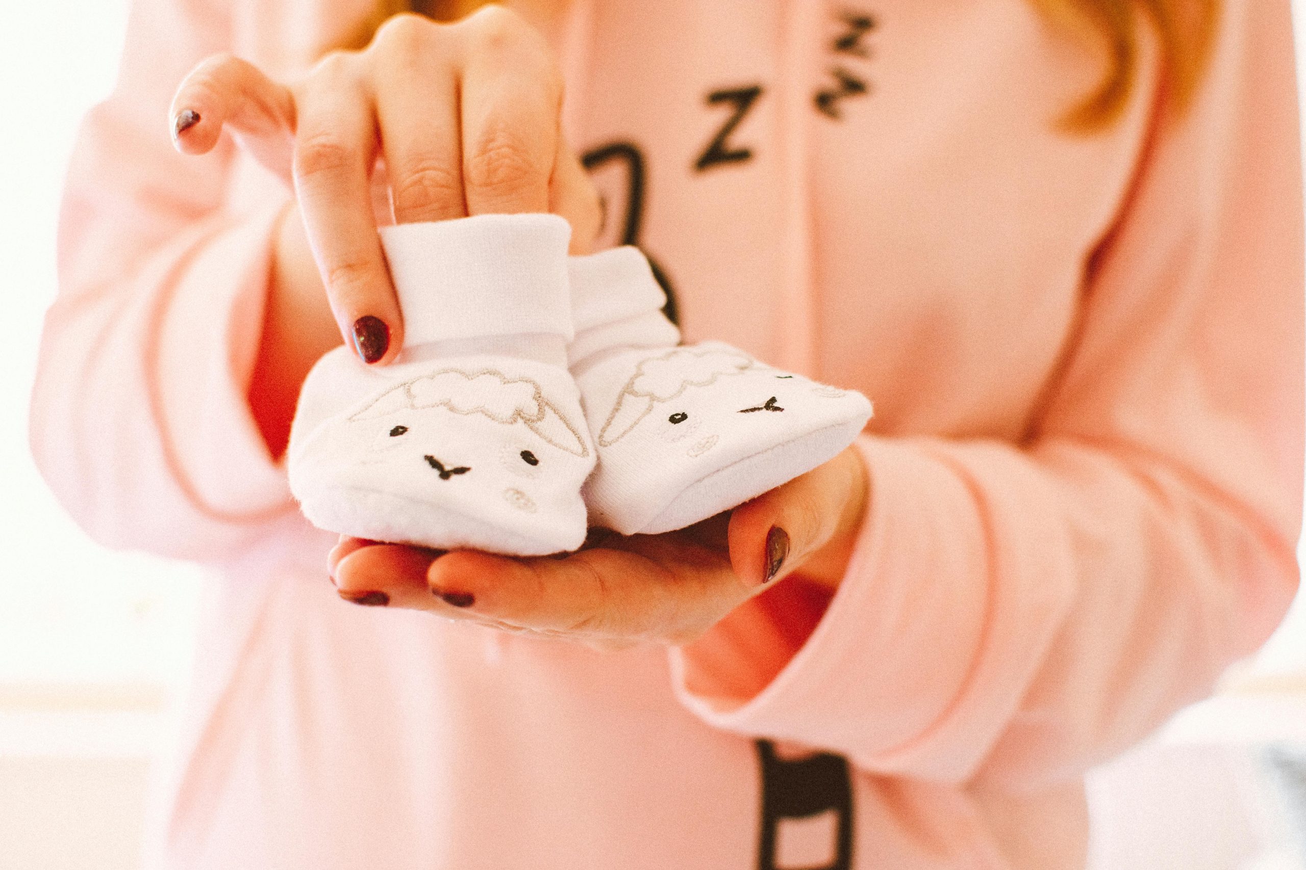 A woman in pink holds cute baby shoes with sheep designs, symbolizing warmth and love.