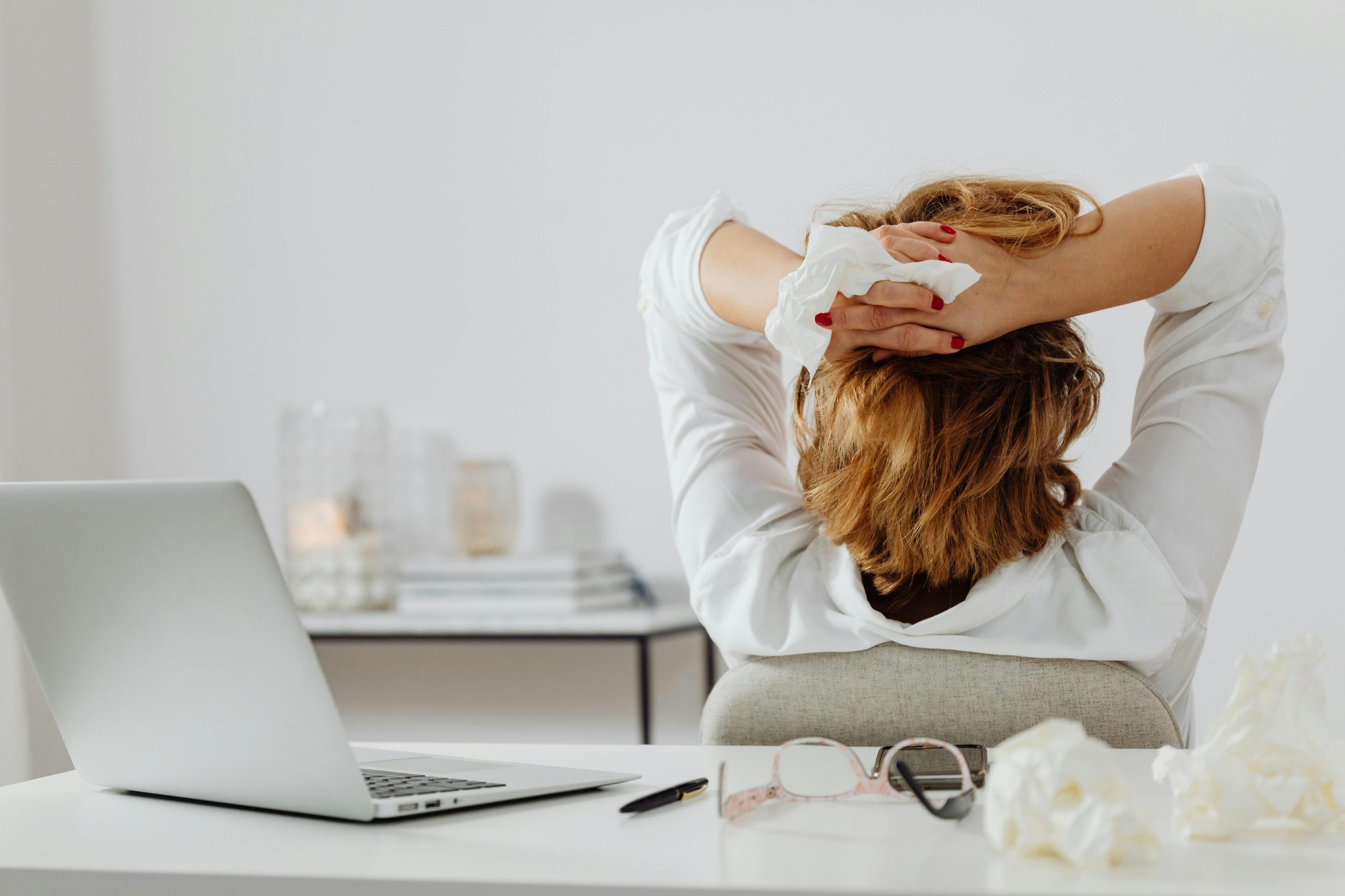 Accompagnement des Dirigeants An adult woman in white takes a break at her desk, holding a tissue, with a laptop and eyeglasses nearby.